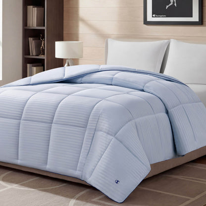 A Blue Cooling Down Alternative Comforter by Champion is displayed on a bed in a modern bedroom with white pillows, a lamp, bookshelf, and framed picture on the wall. The space features neutral decor and soft lighting.