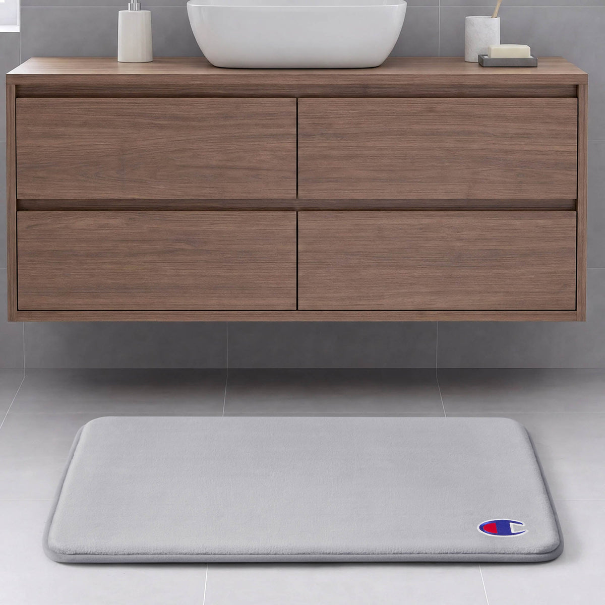 A light gray Champion Logo Patch Memory Foam Bath Mat with a small emblem in the corner rests on a light gray tiled floor in front of a floating wooden bathroom vanity.