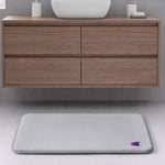 A light gray Champion Logo Patch Memory Foam Bath Mat with a small emblem in the corner rests on a light gray tiled floor in front of a floating wooden bathroom vanity.