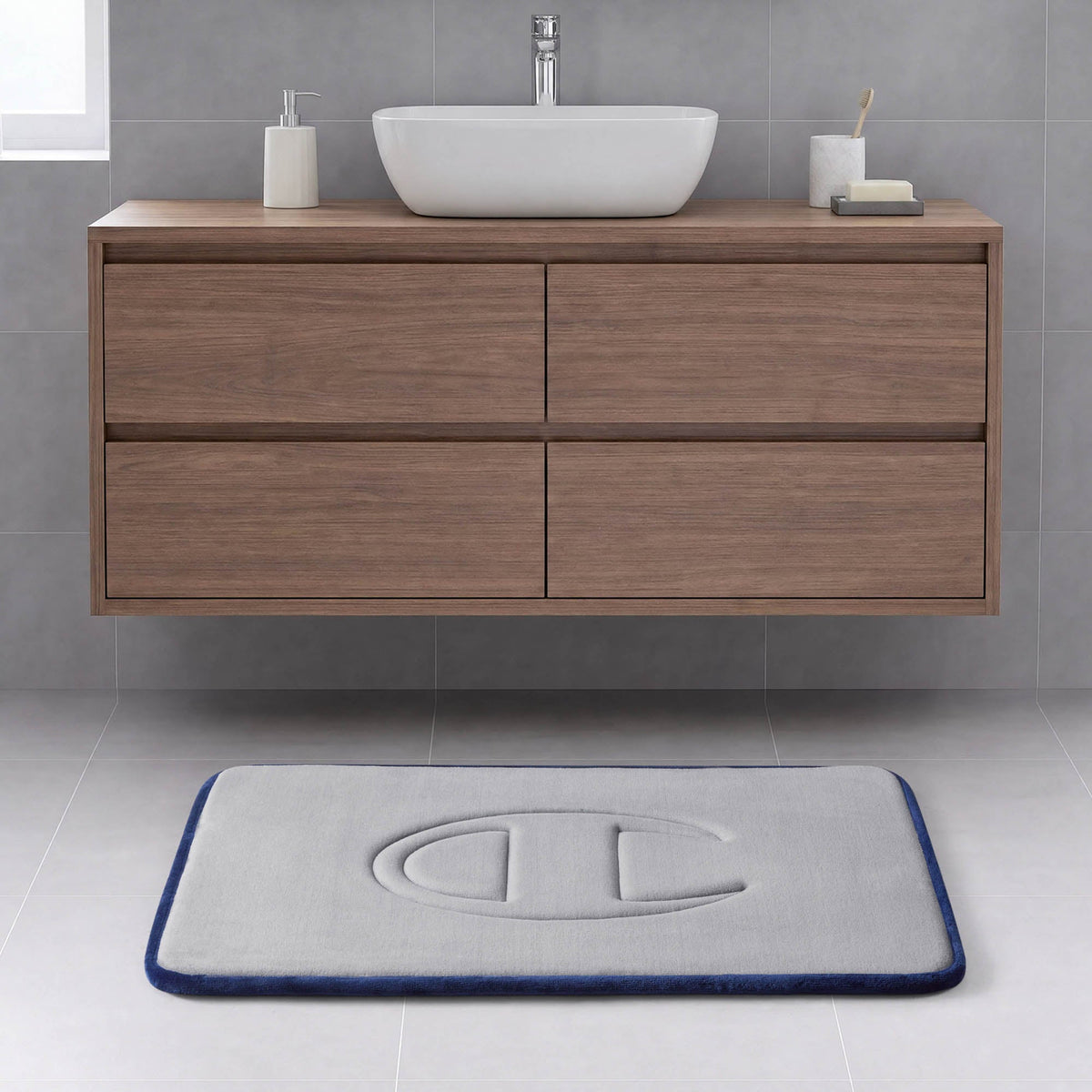A modern bathroom with a floating wooden vanity, a vessel sink, and a rectangular gray Quilted Logo Memory Foam Bath Mat from Champion, featuring an embossed logo, set on a light tile floor.