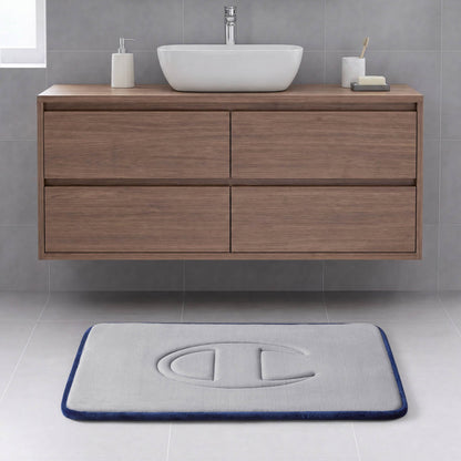 A modern bathroom with a floating wooden vanity, a vessel sink, and a rectangular gray Quilted Logo Memory Foam Bath Mat from Champion, featuring an embossed logo, set on a light tile floor.