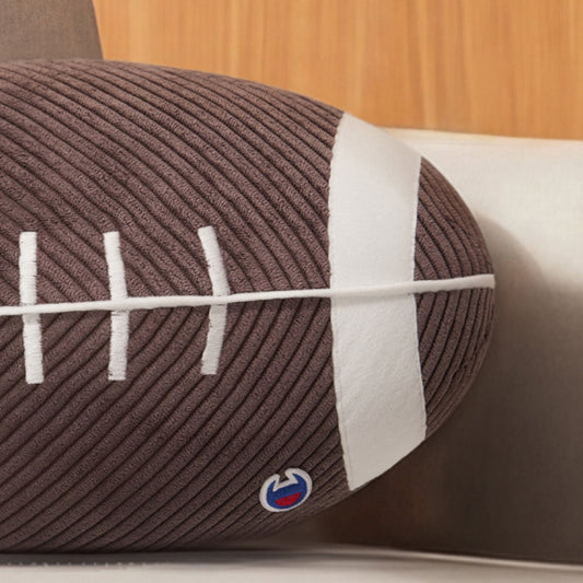A Champion Sport Shaped Pillow, designed like an American football with white laces and stripes, rests on a beige couch.