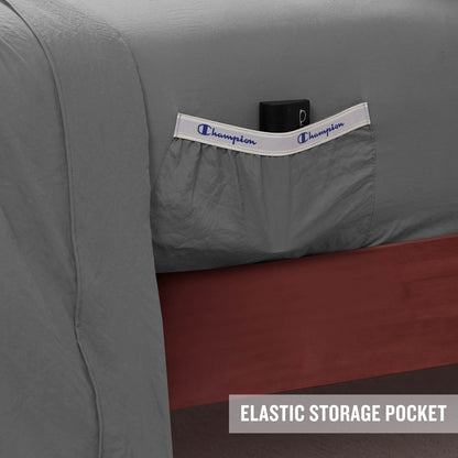 A gray Peak Comfort Microfiber Sheet Set by Champion features an elastic Champion-branded side pocket holding a black item. The sheets are displayed on a wooden bed frame, with an “ELASTIC STORAGE POCKET” label in the corner highlighting the practical design of these deep pocket Champion sheets—ideal for anyone seeking athletic bedding with convenient storage solutions.