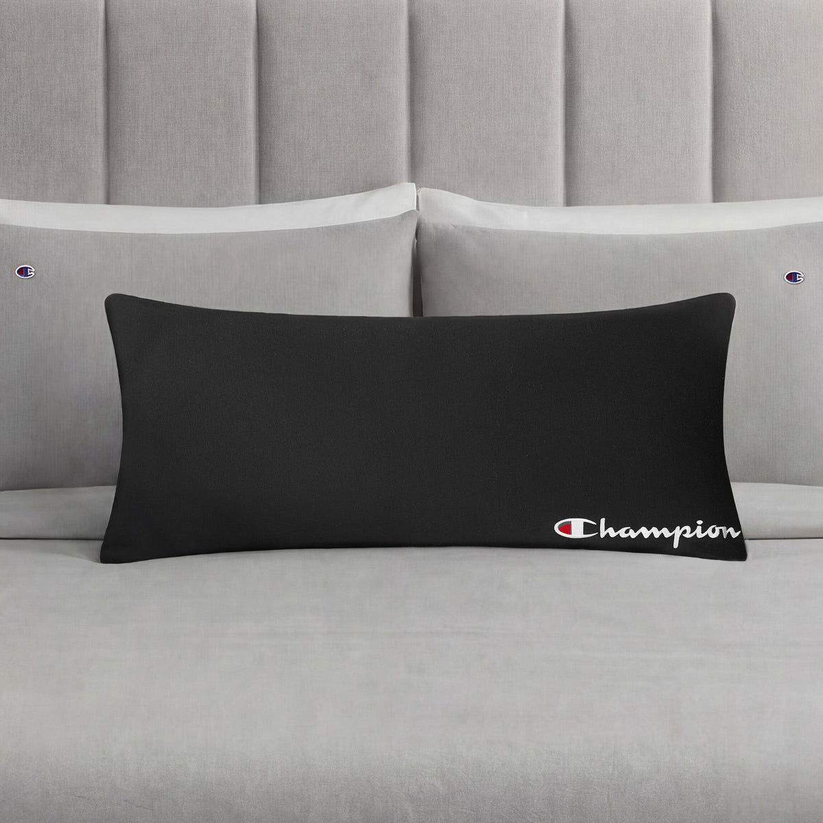 A Champion Jersey Body Pillow in black is displayed on a neatly made bed with light gray bedding and matching pillowcases, each embroidered with small Champion logos, making it the perfect athletic accent pillow for any modern bedroom.