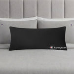 A Champion Jersey Body Pillow in black is displayed on a neatly made bed with light gray bedding and matching pillowcases, each embroidered with small Champion logos, making it the perfect athletic accent pillow for any modern bedroom.