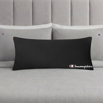 A Champion Jersey Body Pillow in black is displayed on a neatly made bed with light gray bedding and matching pillowcases, each embroidered with small Champion logos, making it the perfect athletic accent pillow for any modern bedroom.