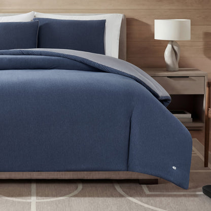A neatly made bed features the Champion Reversible Solid Jersey Comforter Set in blue, with matching pillows arranged against a wooden headboard. On the right side, a white lamp rests on a nightstand, while a patterned beige rug covers the floor. The reversible comforter set adds both style and versatility to the room’s décor.