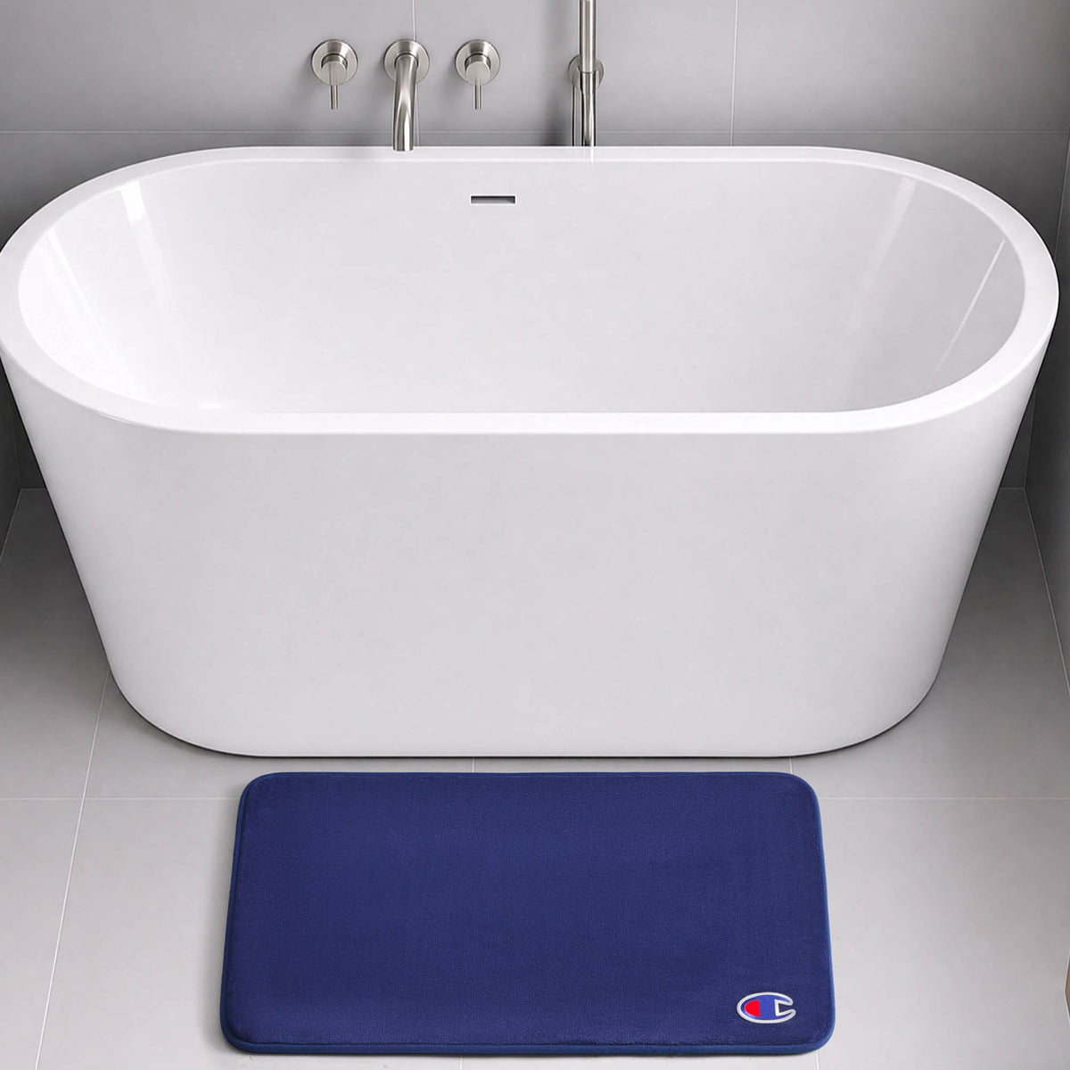 A modern white freestanding bathtub with chrome fixtures sits on a gray tiled floor. In front of the tub, a dark blue Champion Logo Patch Memory Foam Bath Mat with skid-resistant backing and a small logo offers both style and safety.