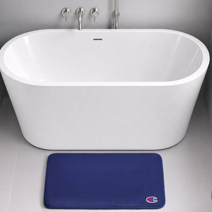 A modern white freestanding bathtub with chrome fixtures sits on a gray tiled floor. In front of the tub, a dark blue Champion Logo Patch Memory Foam Bath Mat with skid-resistant backing and a small logo offers both style and safety.