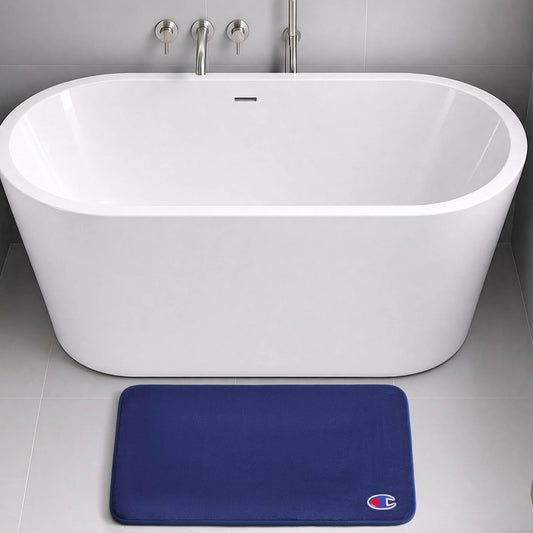 A modern white freestanding bathtub with chrome fixtures sits on a gray tiled floor. In front of the tub, a dark blue Champion Logo Patch Memory Foam Bath Mat with skid-resistant backing and a small logo offers both style and safety.