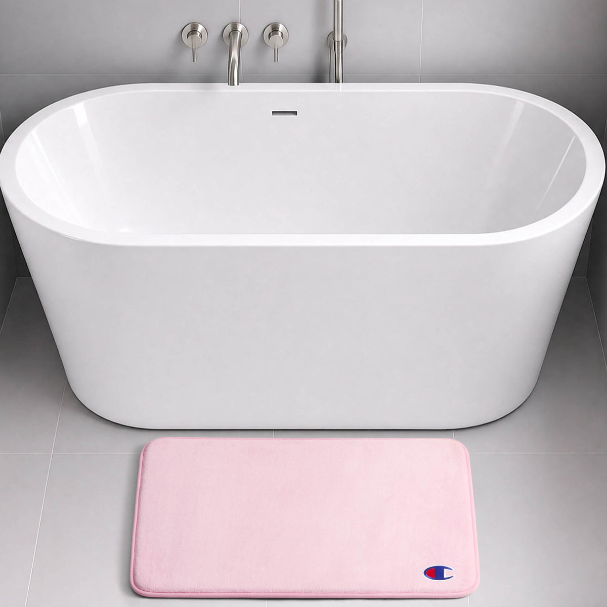 A modern white freestanding bathtub with silver fixtures is set against a gray tiled wall. On the floor in front of the tub lies a light pink rectangular Logo Patch Memory Foam Bath Mat by Champion, featuring a skid-resistant design and adorned with a small blue and red logo.