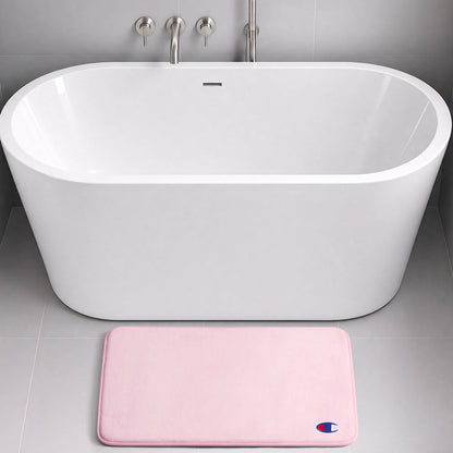 A modern white freestanding bathtub with silver fixtures is set against a gray tiled wall. On the floor in front of the tub lies a light pink rectangular Logo Patch Memory Foam Bath Mat by Champion, featuring a skid-resistant design and adorned with a small blue and red logo.