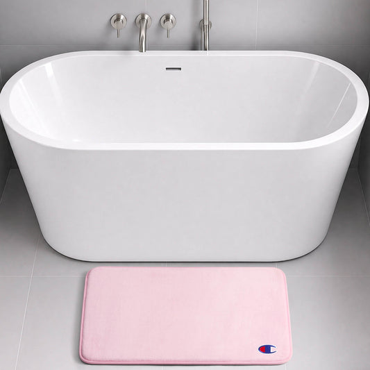 A modern white freestanding bathtub with silver fixtures is set against a gray tiled wall. On the floor in front of the tub lies a light pink rectangular Logo Patch Memory Foam Bath Mat by Champion, featuring a skid-resistant design and adorned with a small blue and red logo.