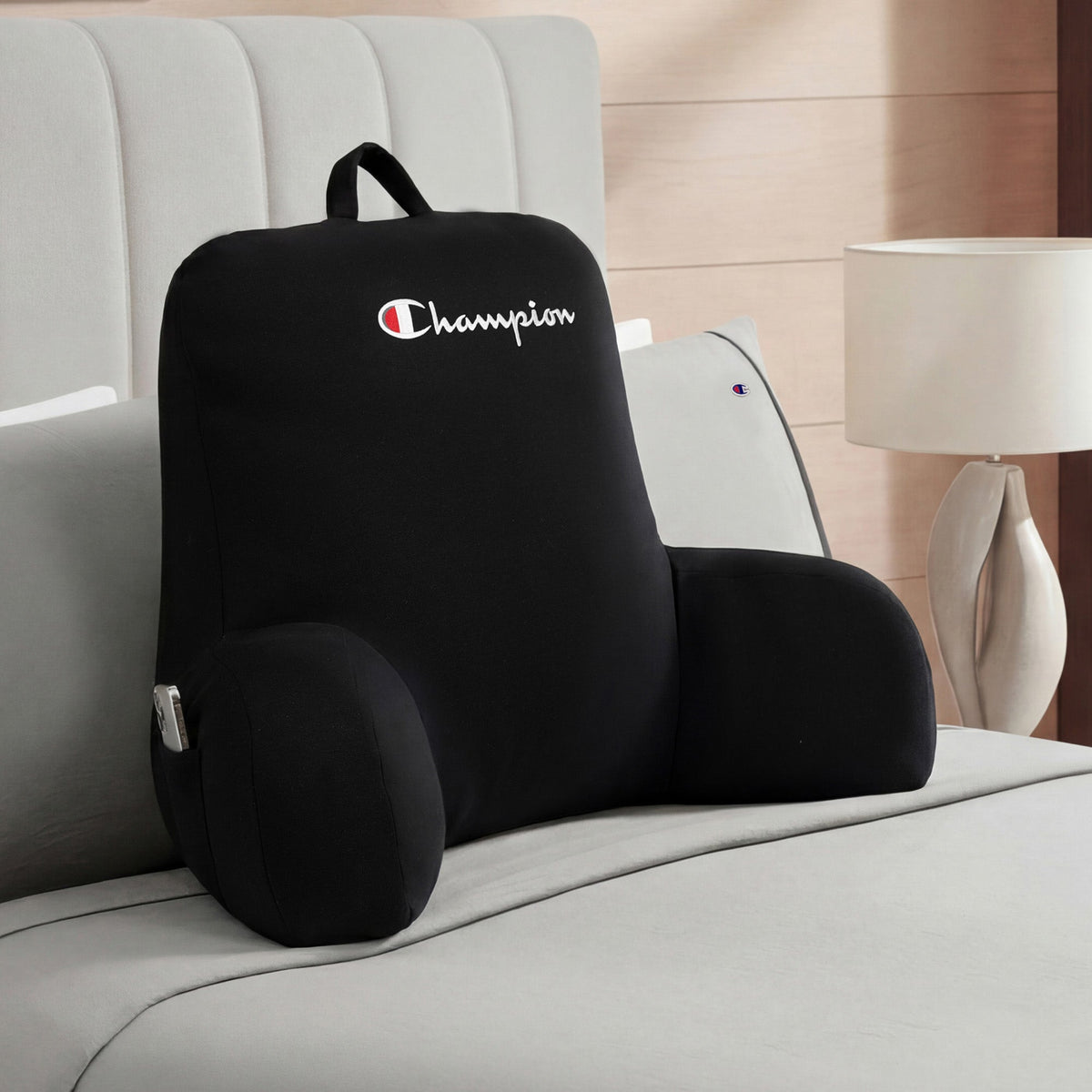 A black Champion Jersey Backrest Pillow with arms rests on a neatly made bed, the iconic Champion logo visible, beside a light-colored lamp on the nightstand in a modern bedroom.
