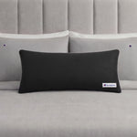 A black rectangular Champion Faux Wool Body Pillow with a small Champion logo rests on a neatly made bed with light gray sheets and two matching pillows, each displaying the Champion brand. The pillow’s plush polyester fill enhances the stylish bedding ensemble with inviting comfort.