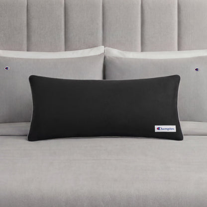 A black rectangular Champion Faux Wool Body Pillow with a small Champion logo rests on a neatly made bed with light gray sheets and two matching pillows, each displaying the Champion brand. The pillow’s plush polyester fill enhances the stylish bedding ensemble with inviting comfort.