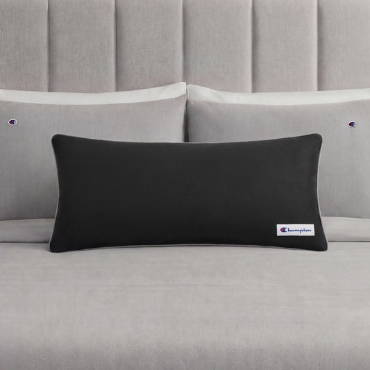 A black rectangular Champion Faux Wool Body Pillow with a small Champion logo rests on a neatly made bed with light gray sheets and two matching pillows, each displaying the Champion brand. The pillow’s plush polyester fill enhances the stylish bedding ensemble with inviting comfort.