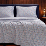 A neatly made bed features the Champion Printed Microfiber Sheet Set in blue and white patterns, paired with two matching pillows. A dark headboard stands against a wood-paneled wall, beside a small table displaying a gold trophy.