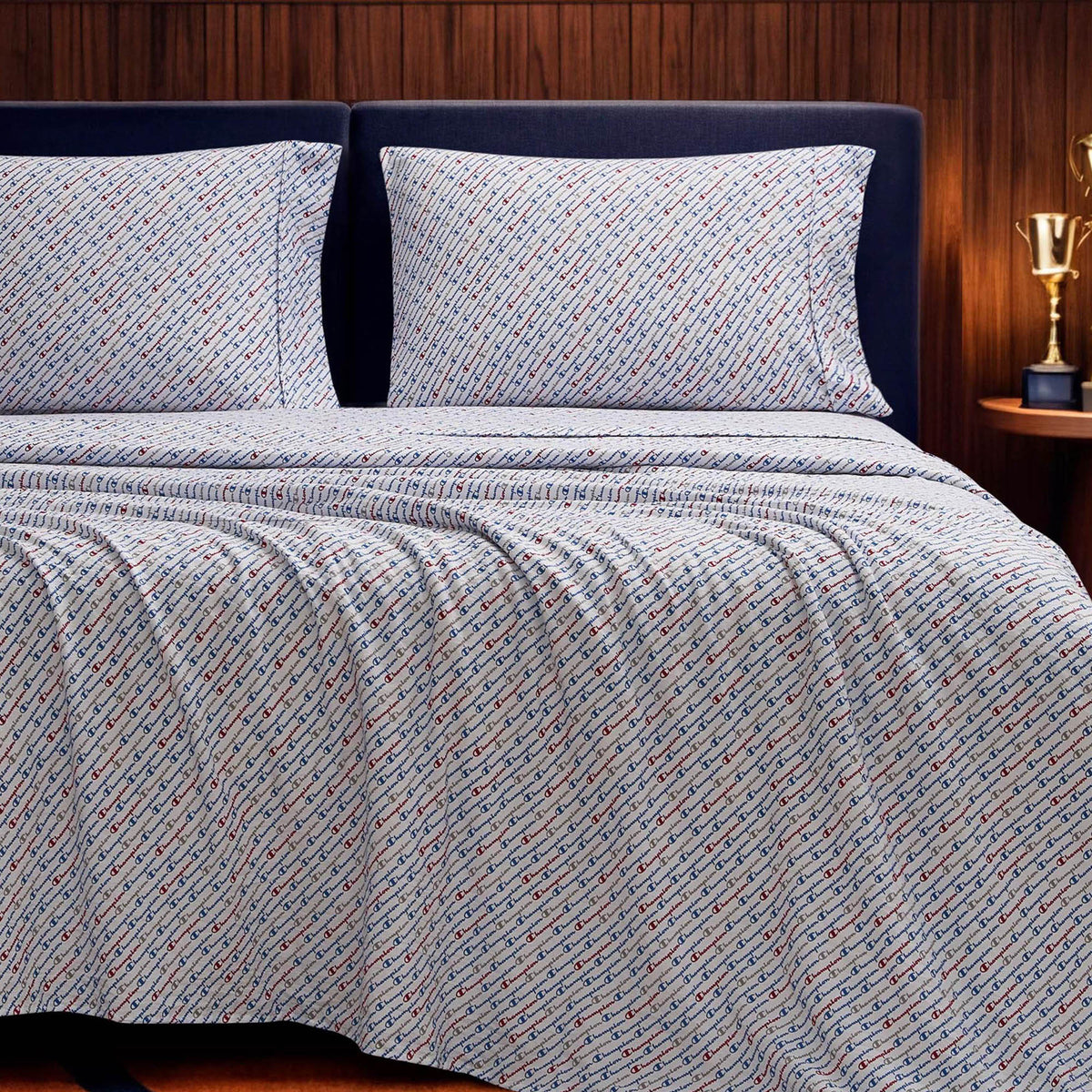 A neatly made bed features the Champion Printed Microfiber Sheet Set in blue and white patterns, paired with two matching pillows. A dark headboard stands against a wood-paneled wall, beside a small table displaying a gold trophy.