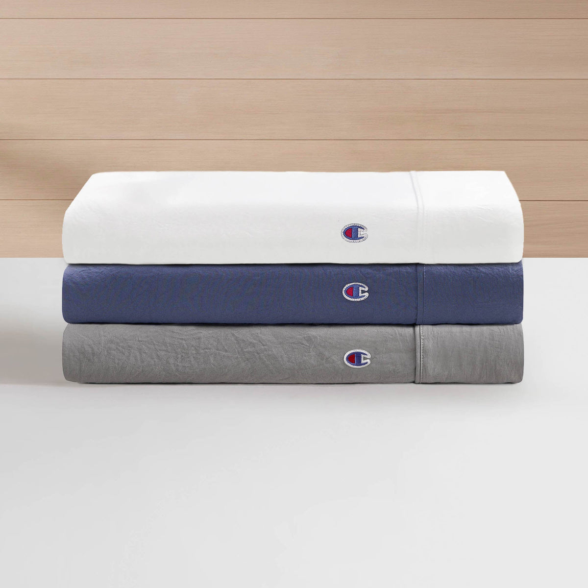 Three folded Champion Embroidery Patch Microfiber Sheet Sets in white, blue, and gray—each featuring a subtle embroidered logo patch—are stacked on a white surface against a light wood background, making for a sleek addition to any Champion bedding collection.