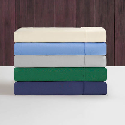 Five folded bedsheets in cream, light blue, light gray, green, and navy—each from the Champion Elastic Logo Microfiber Sheet Set—are stacked on a white surface against a backdrop of vertical dark brown wooden panels.