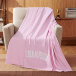 A light pink Champion Jersey with Fleece Backing Throw Blanket featuring “CHAMPION” in white letters is draped over a beige armchair. In the background, a wooden cabinet holds a trophy and a stereo system.