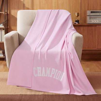 A light pink Champion Jersey with Fleece Backing Throw Blanket featuring “CHAMPION” in white letters is draped over a beige armchair. In the background, a wooden cabinet holds a trophy and a stereo system.