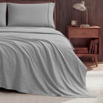 A neatly made bed with light gray Peak Comfort Microfiber Sheet Set by Champion sits next to a wooden nightstand holding a lamp, an alarm clock, and books, set against a dark wood-paneled wall on a light-colored floor.