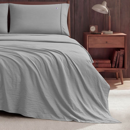 A neatly made bed with light gray Peak Comfort Microfiber Sheet Set by Champion sits next to a wooden nightstand holding a lamp, an alarm clock, and books, set against a dark wood-paneled wall on a light-colored floor.
