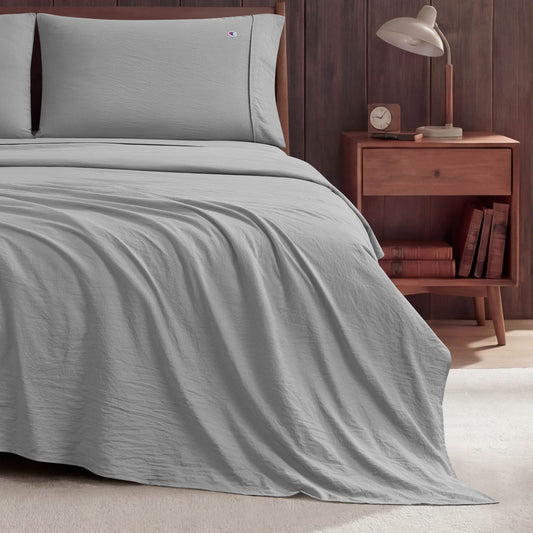 A neatly made bed with light gray Peak Comfort Microfiber Sheet Set by Champion sits next to a wooden nightstand holding a lamp, an alarm clock, and books, set against a dark wood-paneled wall on a light-colored floor.