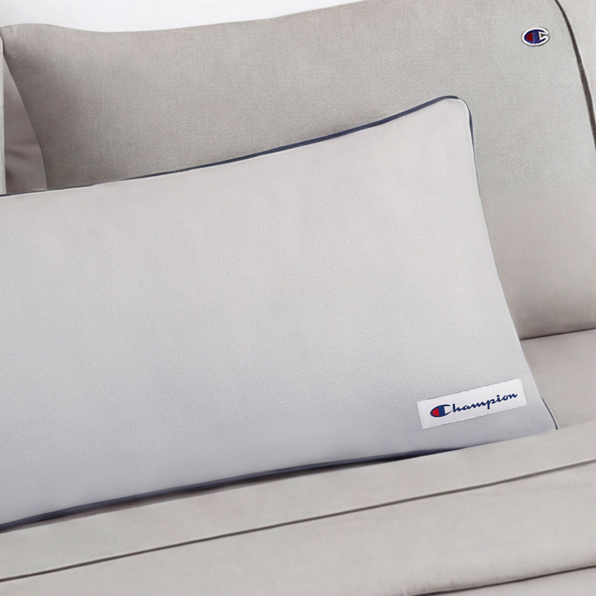 A light gray Faux Wool Body Pillow by Champion, filled with plush polyester and adorned with the Champion logo patch, is elegantly placed on a matching couch or bed for a clean, minimalistic look.