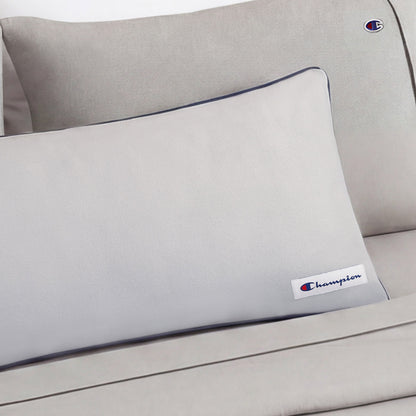 A light gray Faux Wool Body Pillow by Champion, filled with plush polyester and adorned with the Champion logo patch, is elegantly placed on a matching couch or bed for a clean, minimalistic look.