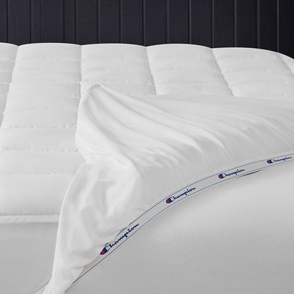 A neatly made bed with a white comforter and sheets, featuring a blue and red Champion logo band along the edge of the top sheet, is complemented by the Champion Ultimate Protection Mattress Pad for added water-resistant protection.