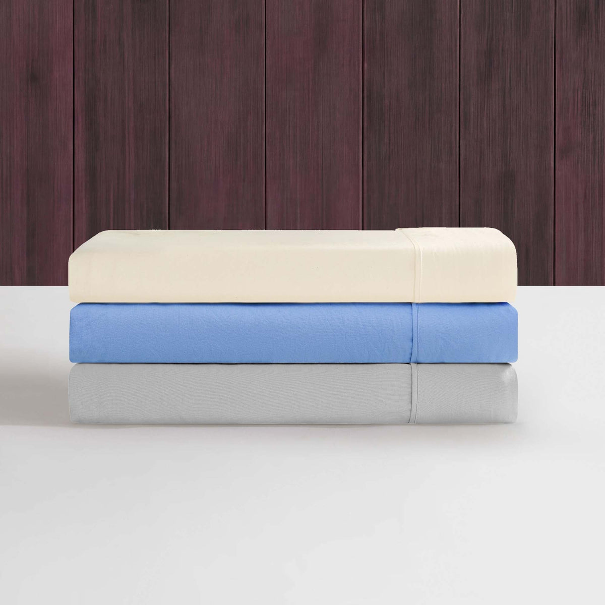 Three neatly folded bedsheets in cream, blue, and gray are stacked on a white surface against a dark wooden wall, highlighting the soft texture of the Champion Elastic Logo Microfiber Sheet Set. Designed by Champion for everyday comfort, each microfiber sheet set features a subtle elastic logo detail that adds a sporty touch.