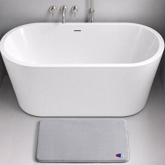 Modern white freestanding bathtub with chrome fixtures, set against gray tiled walls and floor. In front of the tub lies a light gray, skid-resistant Logo Patch Memory Foam Bath Mat from Champion, featuring a small Champion logo.