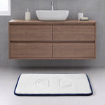 A modern bathroom with a floating wooden vanity, a vessel sink, a soap dish, and the Champion Quilted Logo Memory Foam Bath Mat—with plush cushioning and an embossed logo—accents the light gray tiled floor with its white surface bordered in blue.