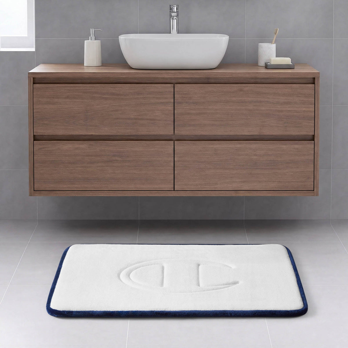 A modern bathroom with a floating wooden vanity, a vessel sink, a soap dish, and the Champion Quilted Logo Memory Foam Bath Mat—with plush cushioning and an embossed logo—accents the light gray tiled floor with its white surface bordered in blue.