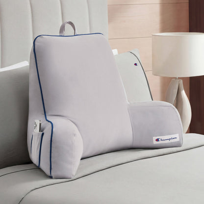 A Champion Faux Wool Backrest Pillow in light gray, featuring supportive arms, blue piping, and convenient side pockets, sits upright on a neatly made bed with gray bedding beside a white lamp on a wooden nightstand.