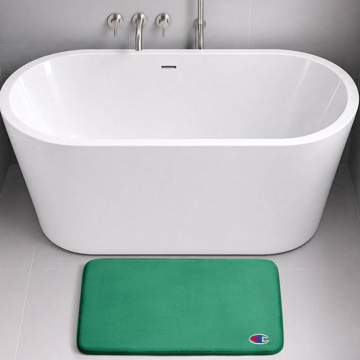 A modern white bathtub with wall-mounted fixtures sits on a gray tile floor. In front of the tub is a green Logo Patch Memory Foam Bath Mat from Champion, featuring a small, colorful C logo in the bottom right corner.