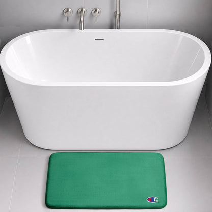 A modern white bathtub with wall-mounted fixtures sits on a gray tile floor. In front of the tub is a green Logo Patch Memory Foam Bath Mat from Champion, featuring a small, colorful C logo in the bottom right corner.