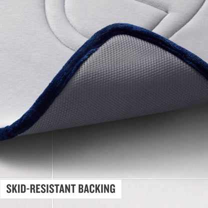Close-up of a gray Champion Quilted Logo Memory Foam Bath Mat with a dark blue border slightly lifted, revealing its textured, skid-resistant backing on a tiled floor. Text in the corner reads, SKID-RESISTANT BACKING.