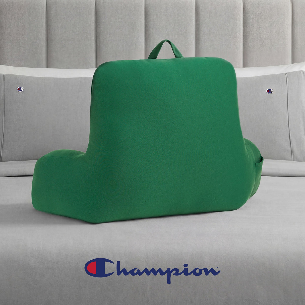 A Champion Jersey Backrest Pillow in green, featuring a convenient handle, rests on a bed with light gray Champion bedding. The iconic Champion logo is visible on the bed next to the backrest pillow, lending a sporty vibe to the cozy scene.