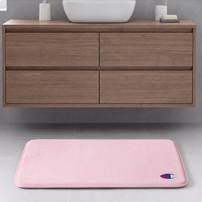 A light pink Champion Logo Patch Memory Foam Bath Mat featuring a small logo in the corner is placed on a gray tile floor in front of a floating wooden bathroom vanity with drawers. This skid-resistant memory foam mat combines style and safety for your space.