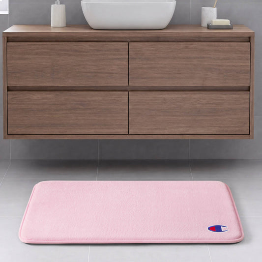 A light pink Champion Logo Patch Memory Foam Bath Mat featuring a small logo in the corner is placed on a gray tile floor in front of a floating wooden bathroom vanity with drawers. This skid-resistant memory foam mat combines style and safety for your space.