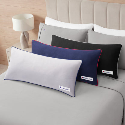 Three rectangular Champion Faux Wool Body Pillows in white, navy, and black, each featuring colored piping and the classic Champion logo, are arranged on a neatly made bed with gray bedding next to a white lamp on a wooden nightstand.