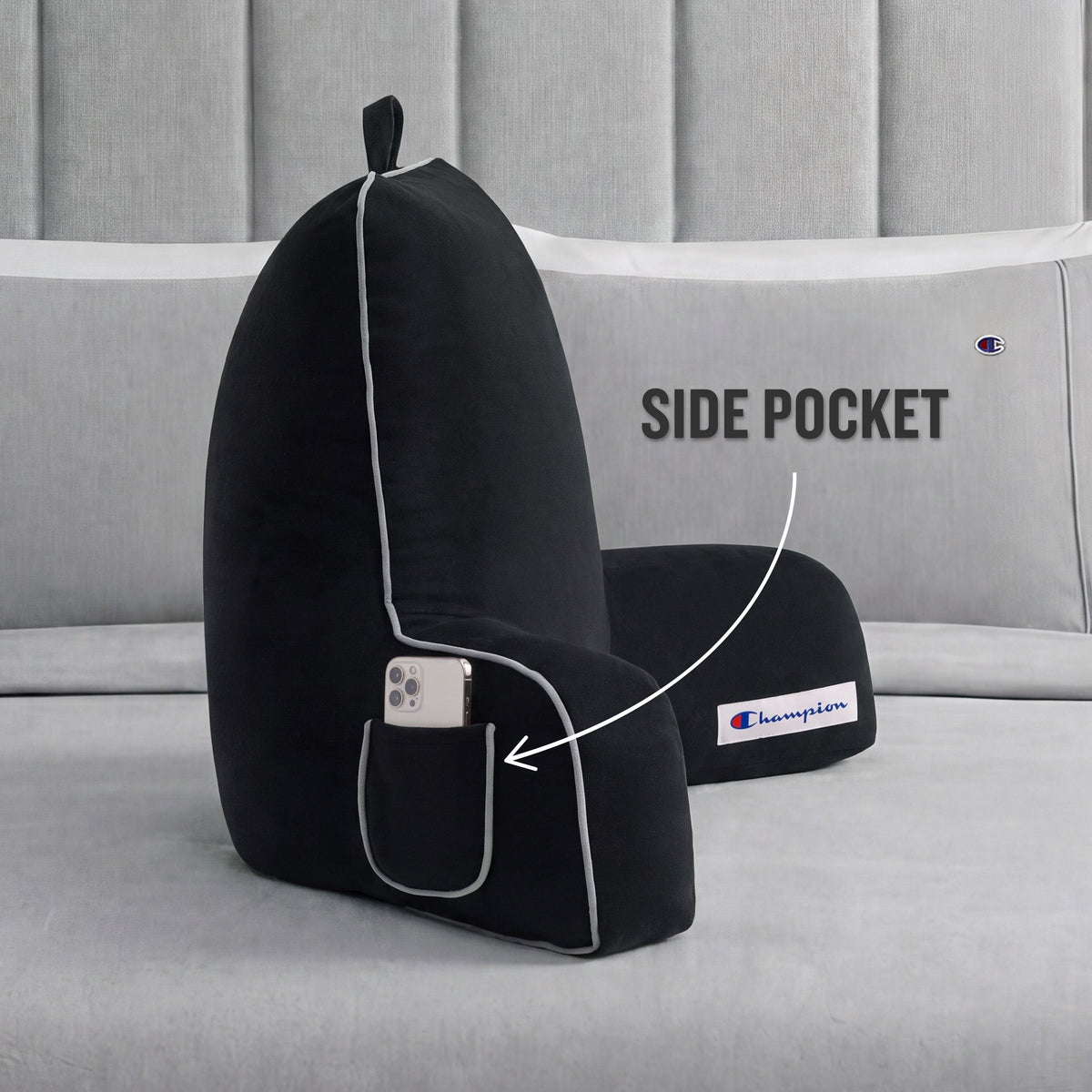 A black Champion Faux Wool Backrest Pillow with supportive arms rests on a bed. An arrow highlights the side pocket containing a smartphone, with text that reads, SIDE POCKET.