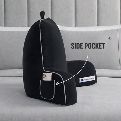 A black Champion Faux Wool Backrest Pillow with supportive arms rests on a bed. An arrow highlights the side pocket containing a smartphone, with text that reads, SIDE POCKET.