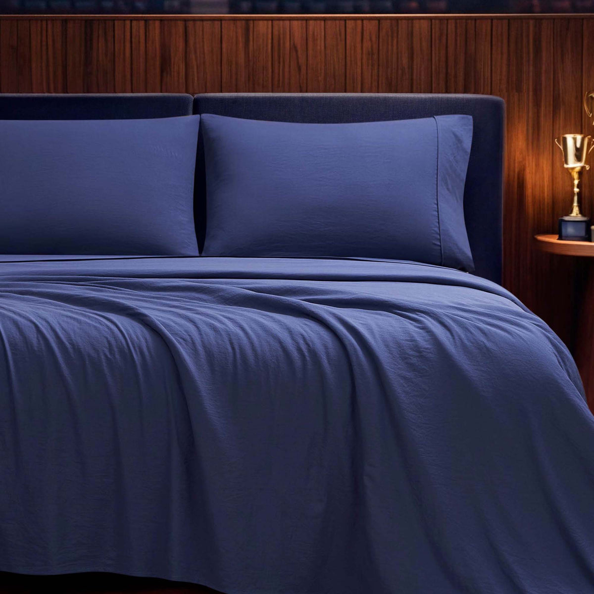 A neatly made bed features the Champion Elastic Logo Microfiber Sheet Set in navy blue, complete with matching pillowcases, set against a wooden headboard. On the small round nightstand sits a gold trophy-shaped lamp.