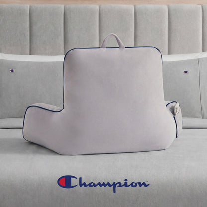 A Champion Faux Wool Backrest Pillow in light gray, featuring supportive arms, a handle, and a side pocket, is placed on a gray bed. The bedspread displays the Champion logo in blue and red, and matching Champion-branded pillows complete the bedding ensemble.