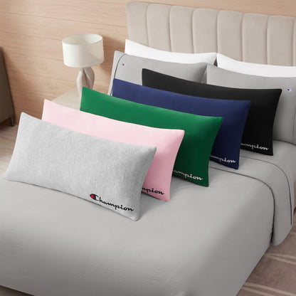 Five Champion Jersey Body Pillows in gray, pink, green, blue, and black—including a cozy jersey knit style—are neatly arranged on a bed with light gray bedding for a sporty accent. A bedside table with a lamp sits next to the bed.